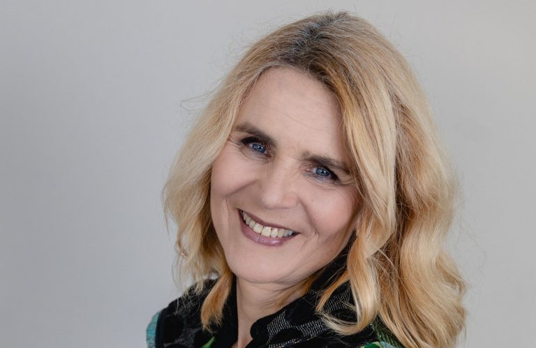 Selbstportrait Blonde Frau mit lächelndem Gesicht, trägt einen bunten, gemusterten Pullover.